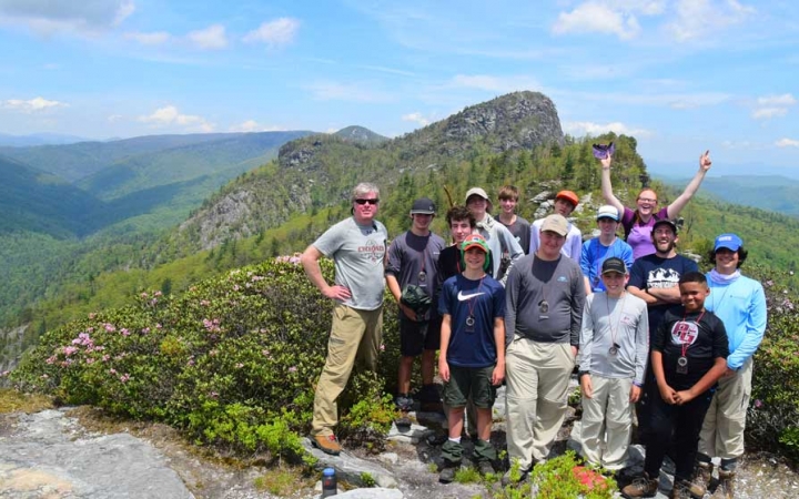 A group of students poses for a photo at high elevation. There is a vast, tree-covered mountainous landscape behind them.